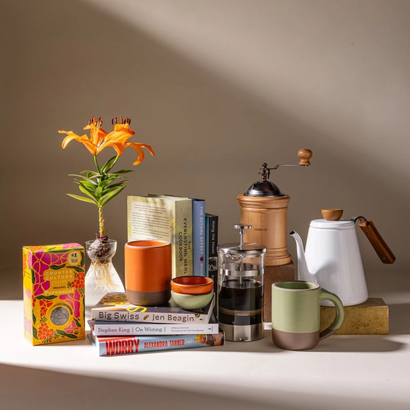 A lineup of various goods all together including ceramic mugs, a clear bulb vase, a stack of books, some coffee accessories like a french press, and a box of coffee.