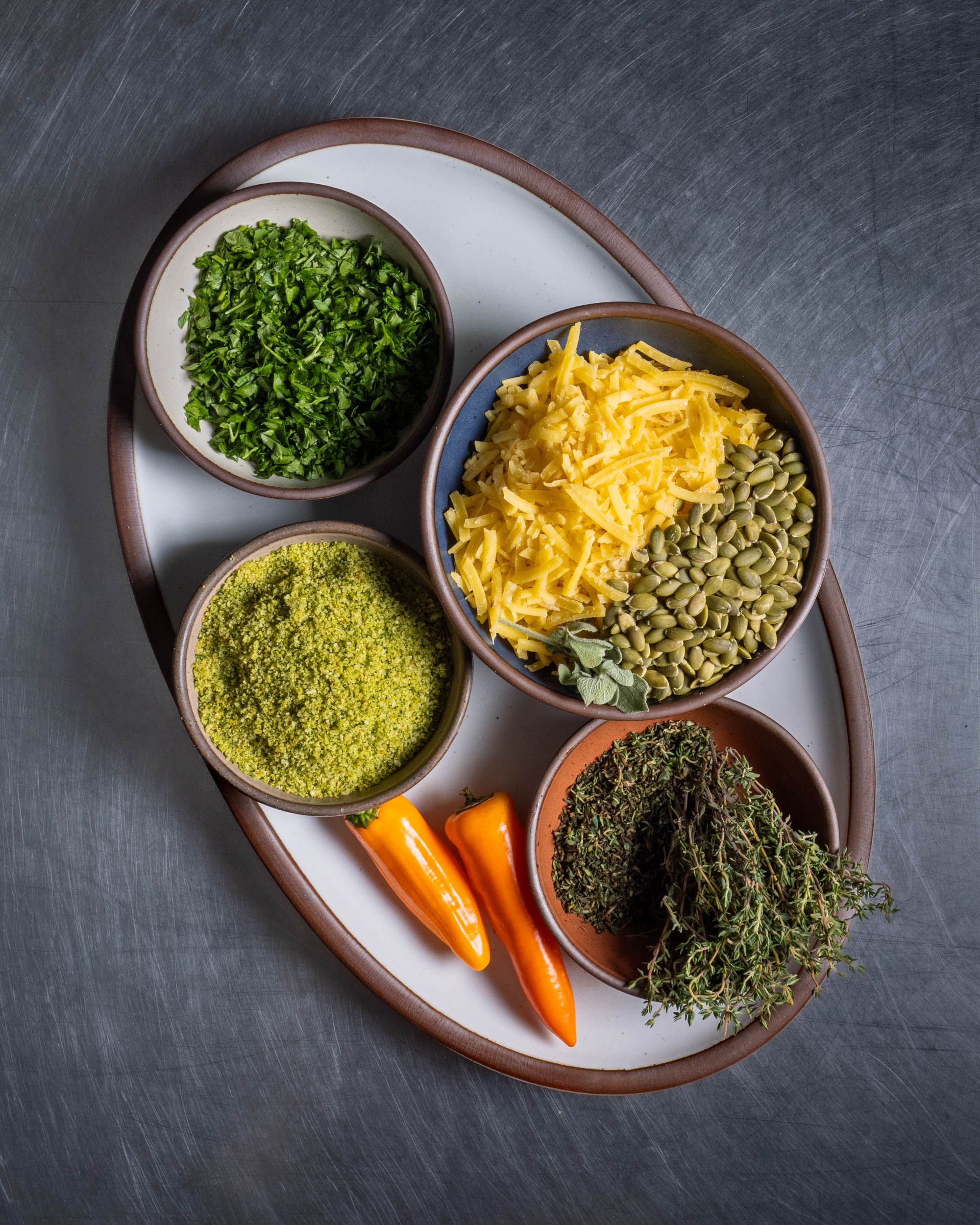 Ceramic plates and bowls hold ingredients for mac and cheese including shredded cheese, peppers, and herbs.