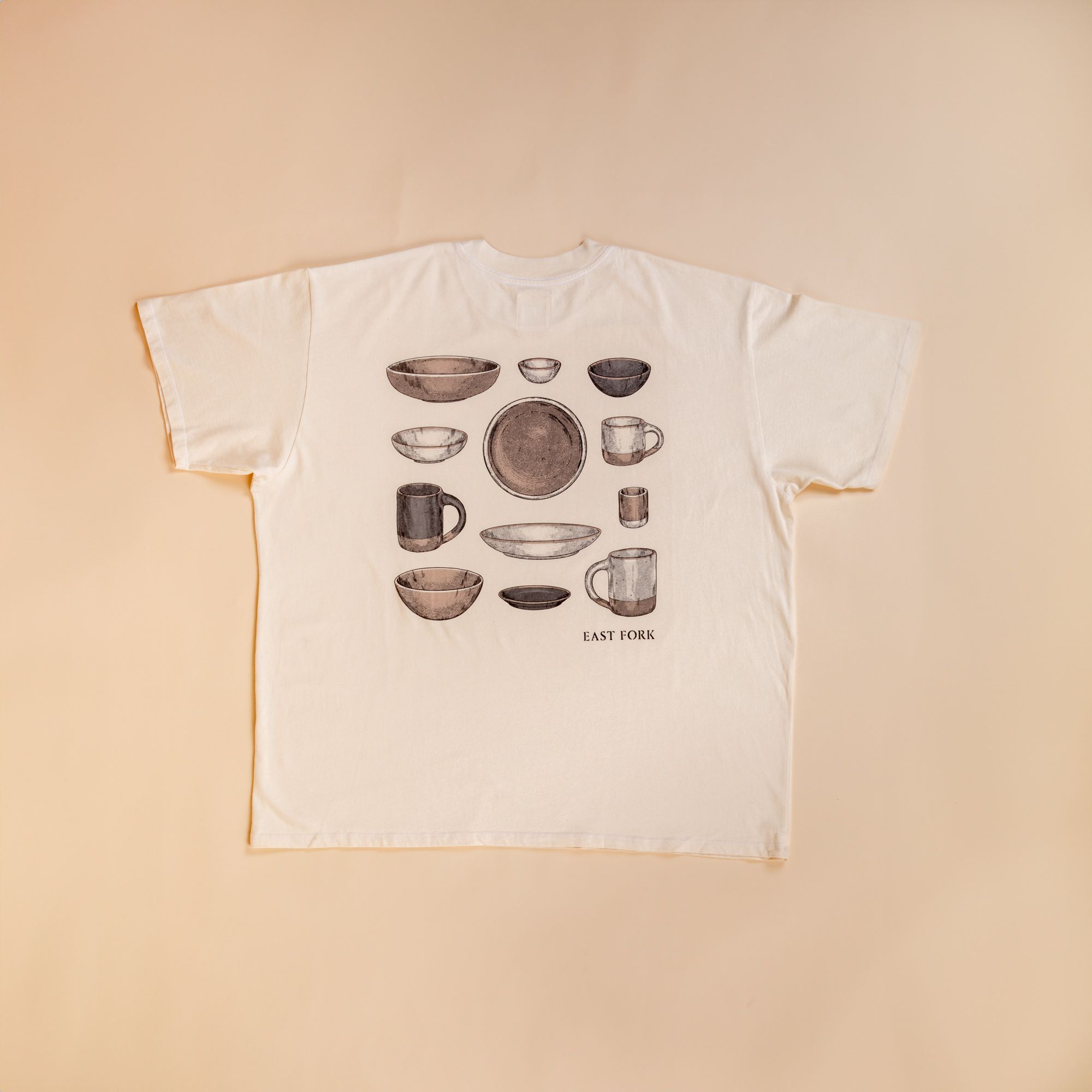 Flat lay of the back of a t-shirt in cream against a cream background. The back has an illustration of various mugs, plates, and bowls all in neutral colors.