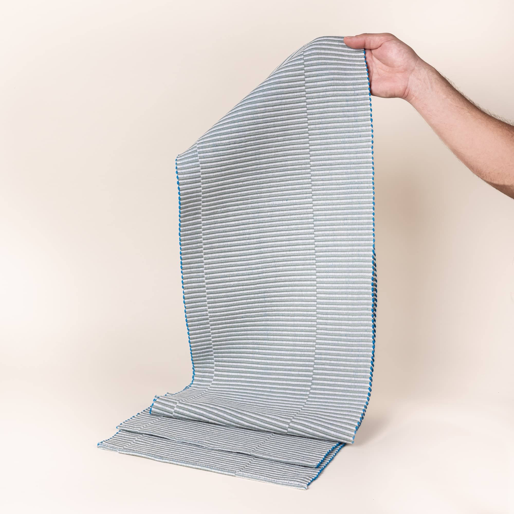 A hand holds up a table runner with offset stripe design that is hand woven in sage and blue colors.