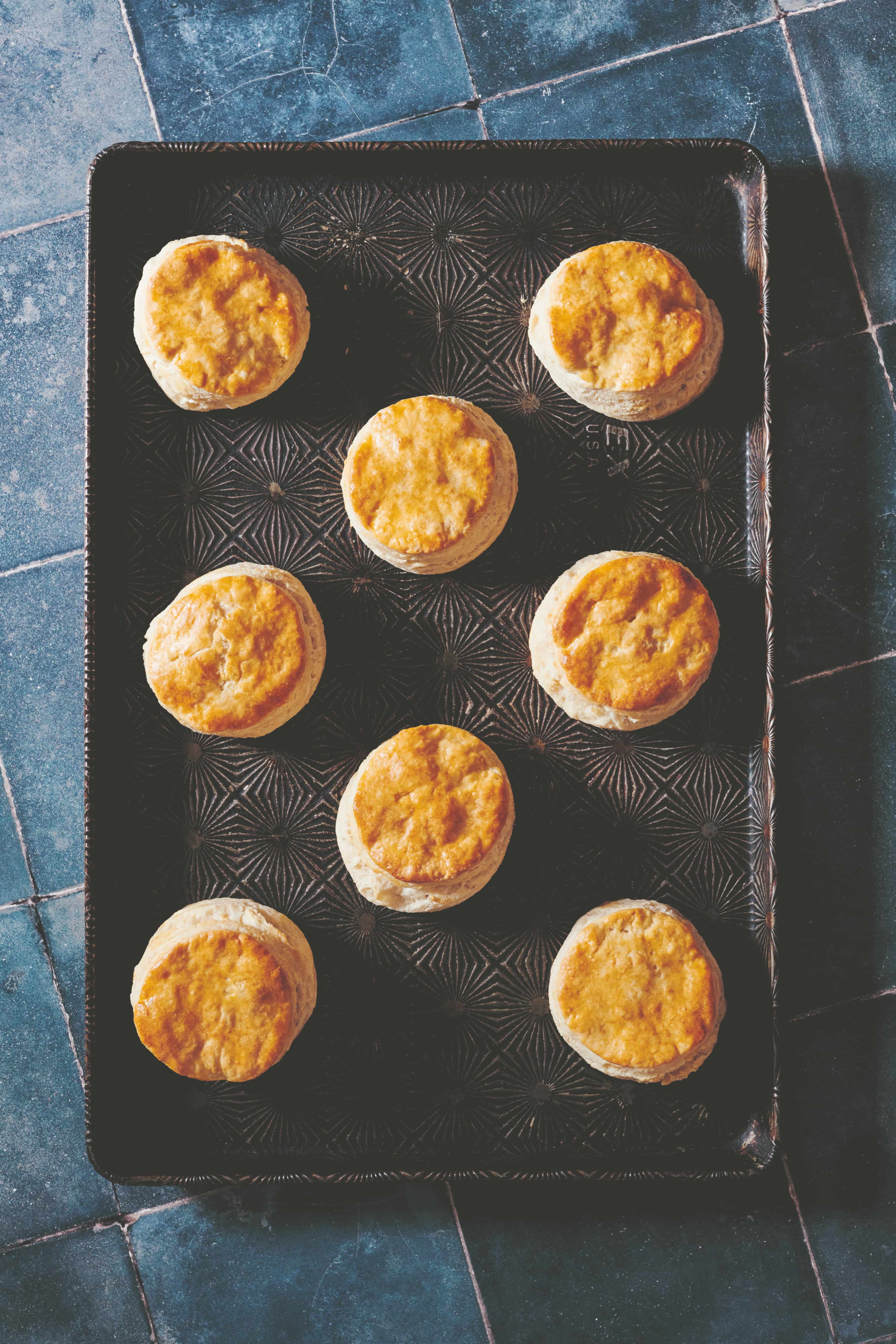 Overhead view of golden biscuits on an oven tray against a blue tile countertop.