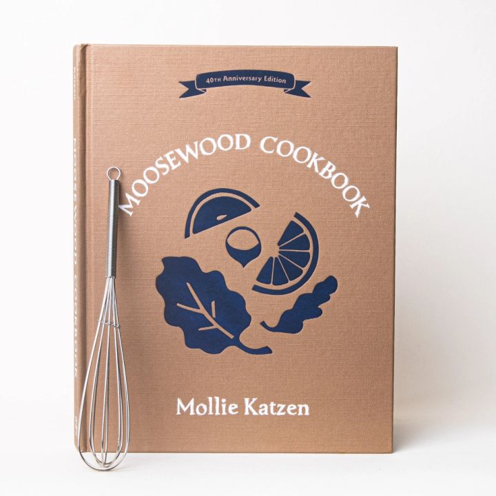 A mini whisk in front of a book with a kraft brown color and silhouette illustrations of leaves and fruit slices with a title that reads "Moosewood Cookbook"