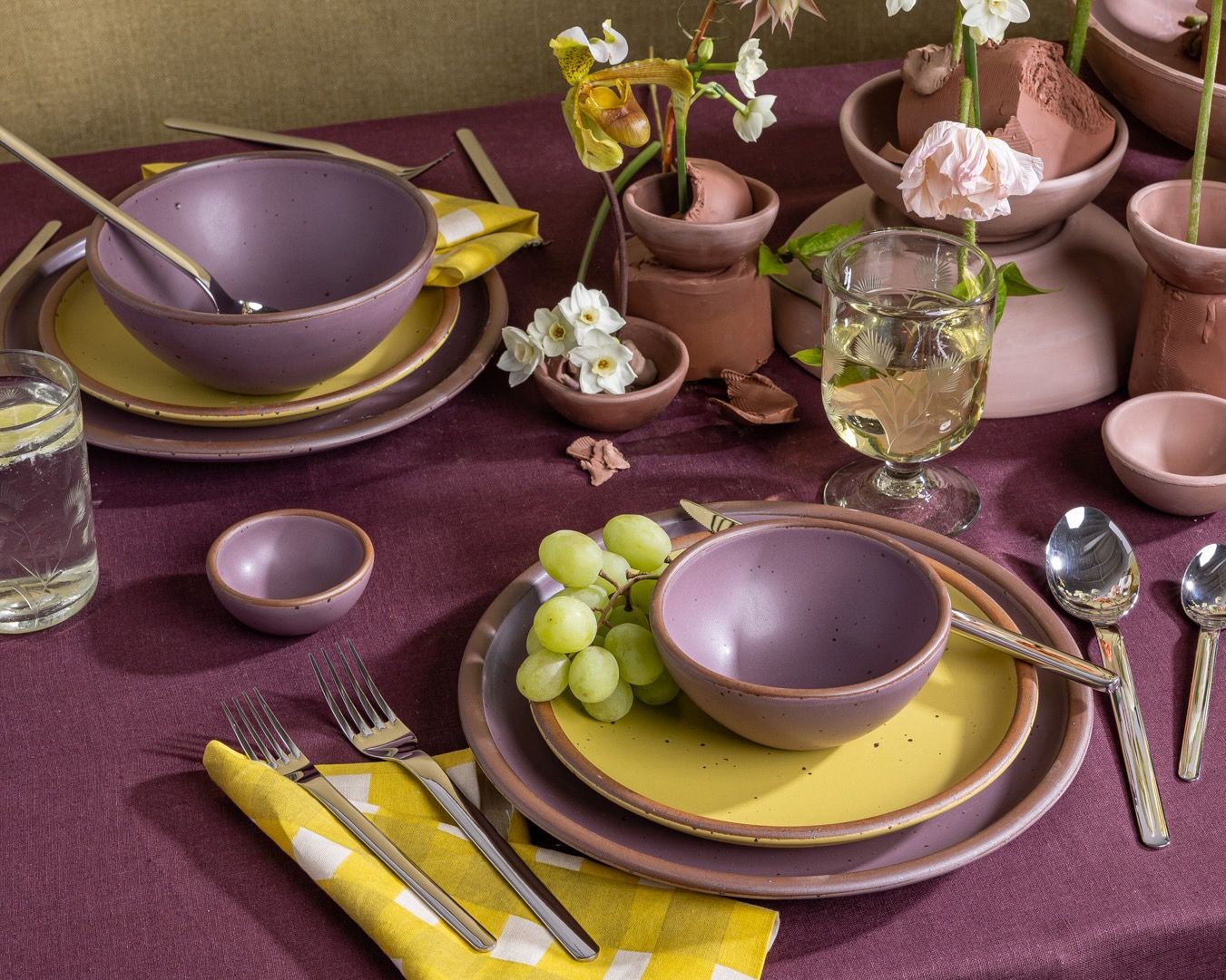 A table featuring two place settings with zesty yellow and soft purple ceramic plates and bowls, with silverware, flowers, and colorful yellow and purple textiles.