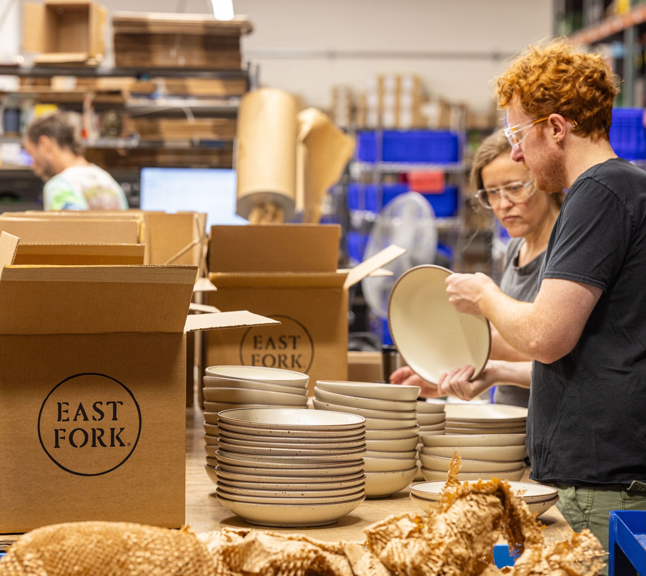 In a factory setting, people are examining ceramic plates and large shallow bowls to pack into large cardboard boxes that say "East Fork"