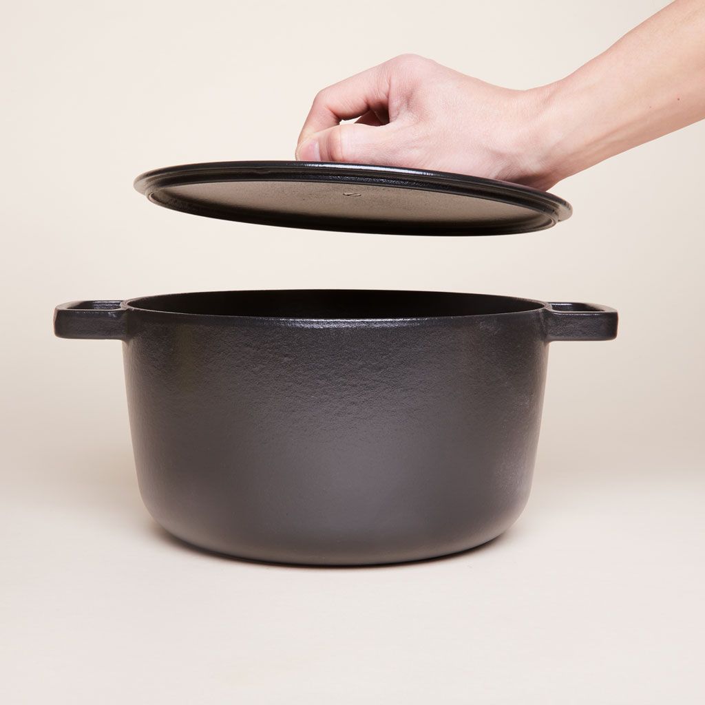 A hand lifts the lid from a lidded black pot with handles made of cast iron