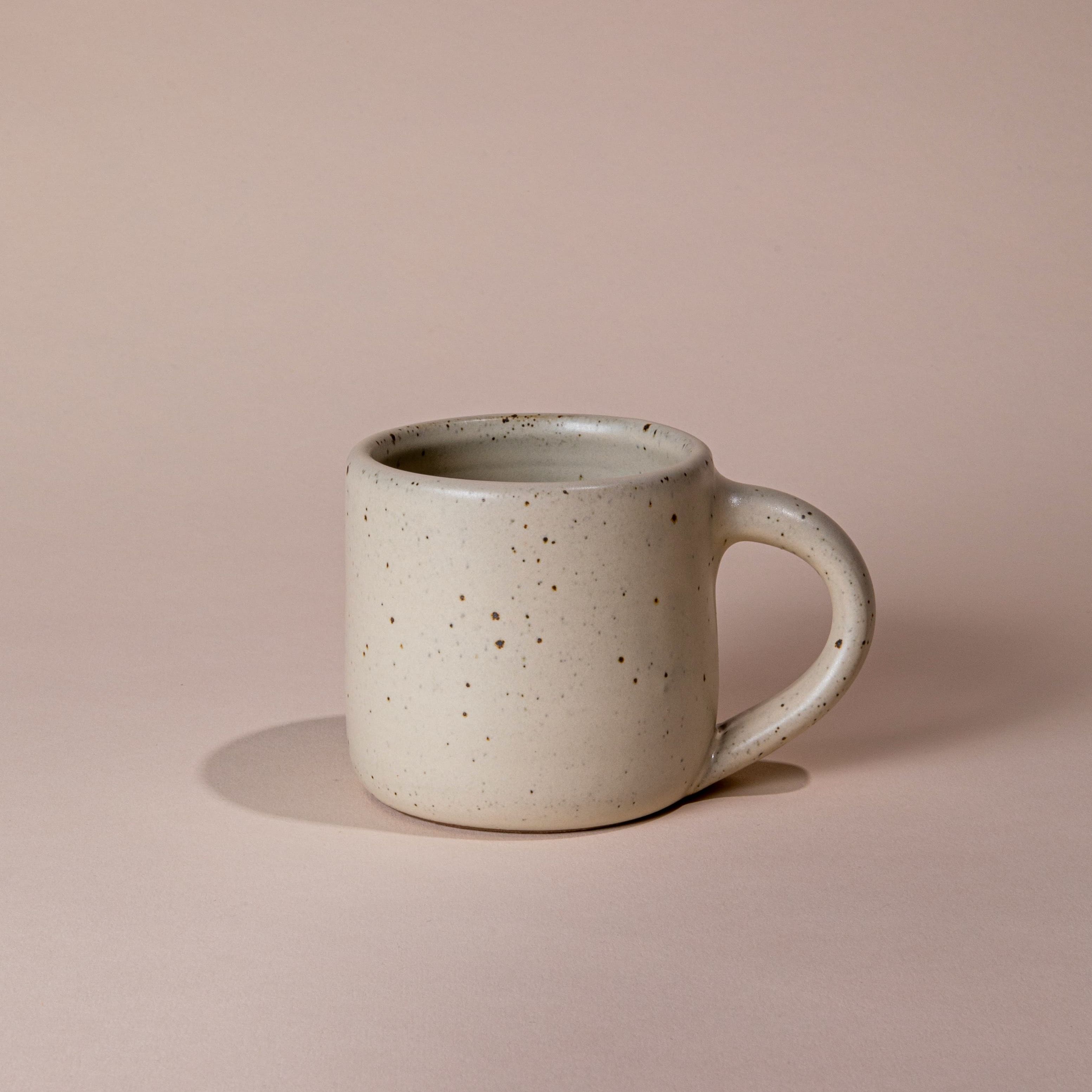 A medium sized ceramic mug with handle in a warm, tan-toned, off-white color featuring iron speckles