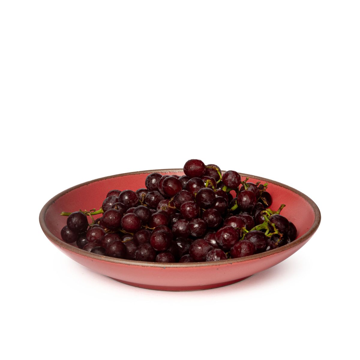 Grapes fill a large ceramic plate with a curved bowl edge in a bold red color featuring iron speckles and an unglazed rim.