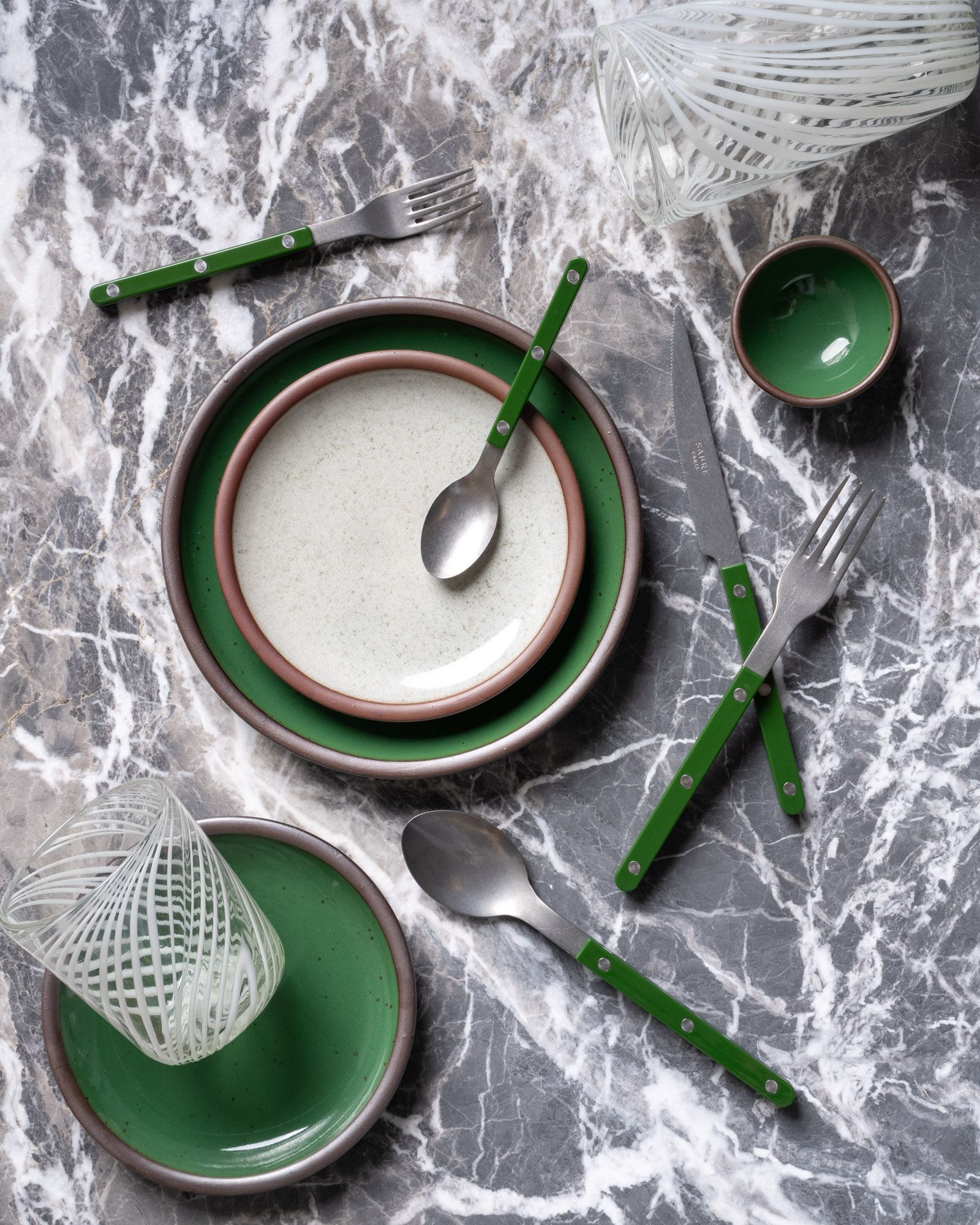 Stacked glossy green and reflective icy white ceramic plates with unglazed rims, green-handled flatware, and clear patterned glasses on a gray marble surface.