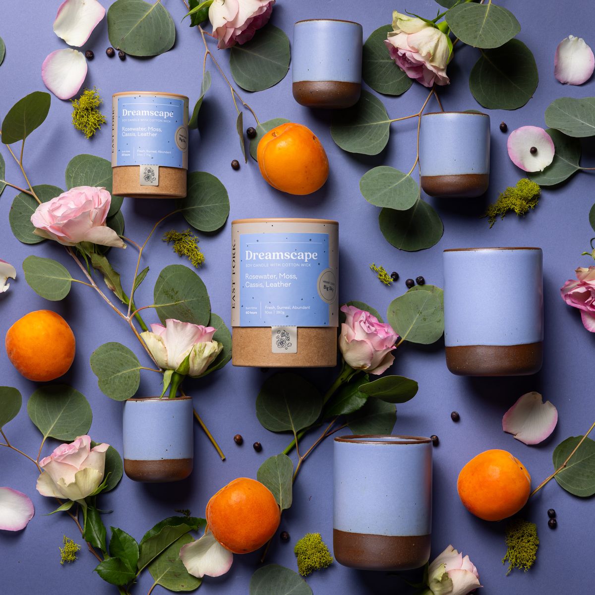 An artful layout of The Candle in Dreamscape - centered is a cardboard packaging tube with the candle's name, surrounded by small and large ceramic candles in a cylindrical vessel in a periwinkle color, surrounded by roses, fruits, and greenery