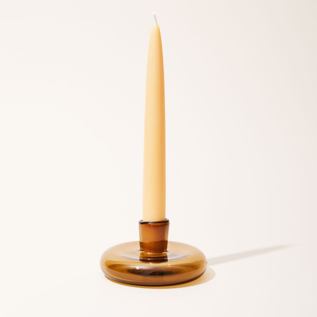 A tall beige candle stands in a clear brown candle holder made of a disc-like base with a small cup at center.