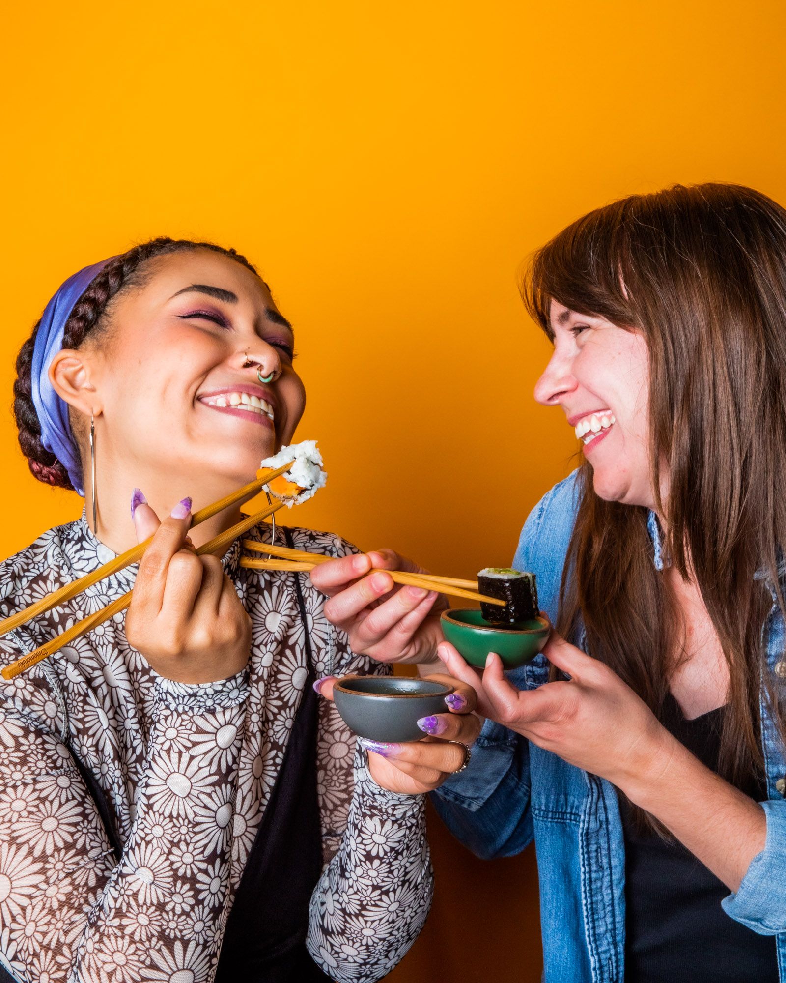 Two people, smiling at each other against a yellow backdrop, holding chopsticks with sushi in one hand and a little bowl in grey and green respectively.
