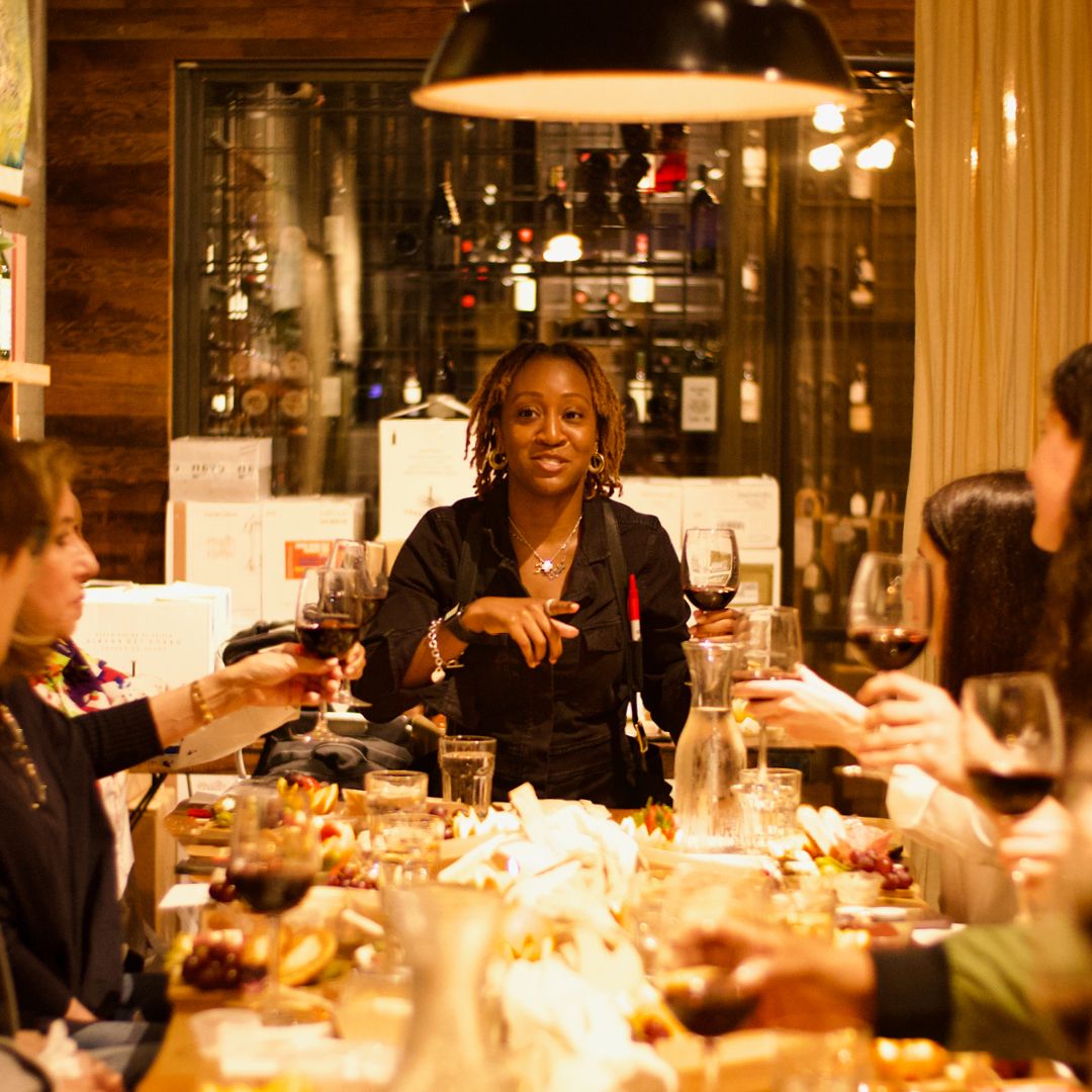 In a restaurant setting, a group of people are gathered around a table filled with glasses, plates, and food. The head of the table is standing and presenting.