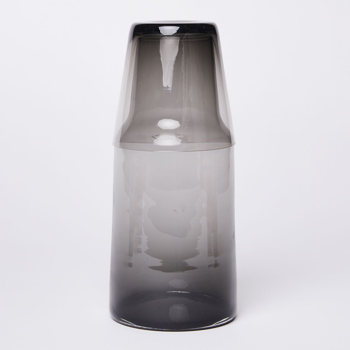 Glass carafe tapering at the top with a dark black bottom getting lighter grey towards the top with a black to grey glass stacked upside down on top of the carafe 