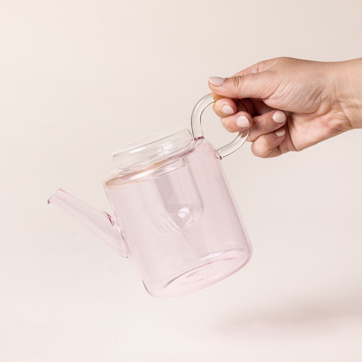 A hand holds a modern glass teapot in a faint pink color with a strainer inside