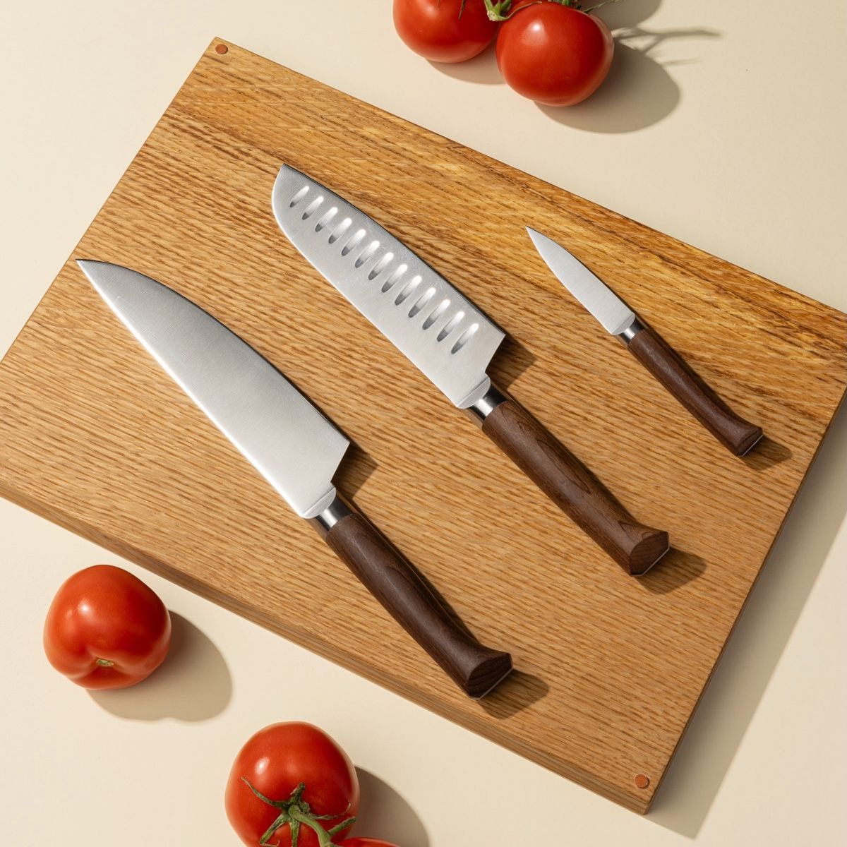 Three different kitchen knives lined up on a wooden cutting board with tomatoes surrounding. The knives are a chef's knife, a santoku knife, and a paring knife.