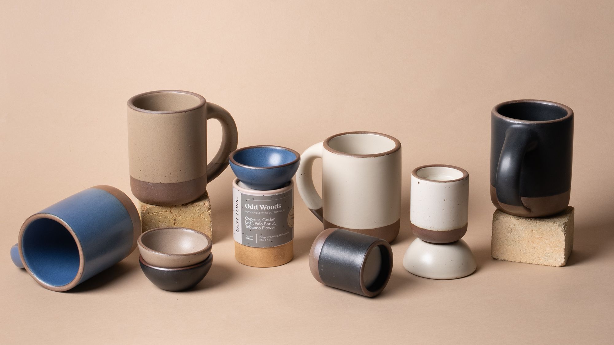 A collection of East Fork mugs, bowls, and candles in neutral and blue glazes arranged on a beige background with textured blocks for height variation.