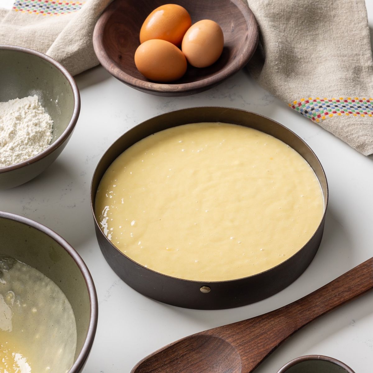 A round black metal cake pan filled with cake batter on a countertop. Surrounded by a bowl of batter, wooden spoon, bowl of eggs, and a kitchen towel.