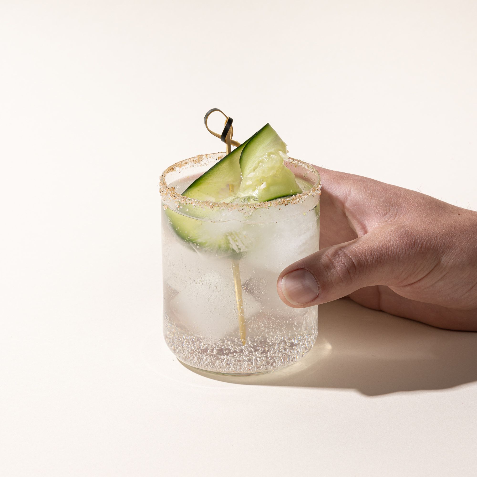 A hand holds a straight walled short glass cup with a clear cocktail and cucumber garnish.
