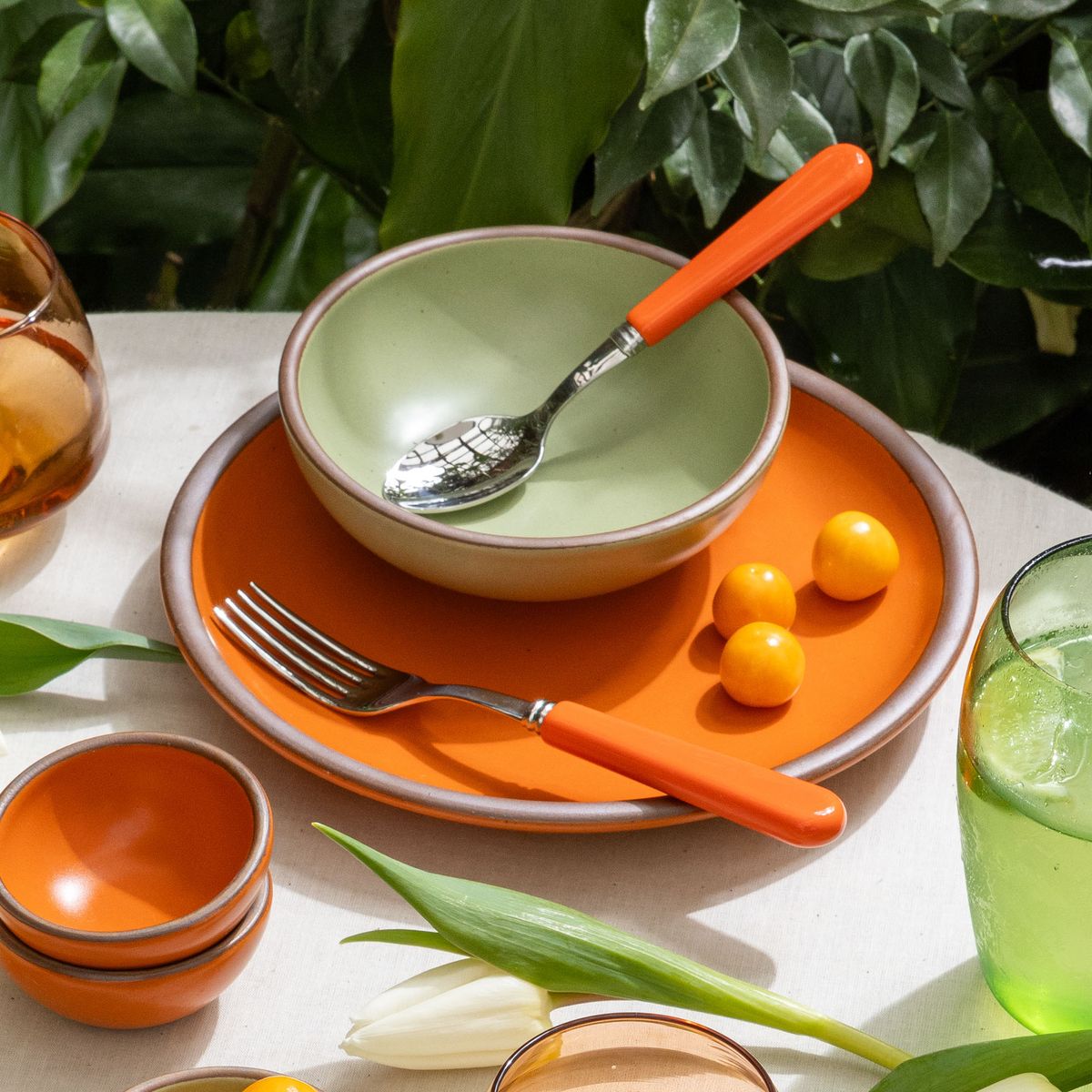 A table setting featuring a bold orange plate and tiny bowls, a sage green shallow bowl, and bold orange handled fork and spoon.
