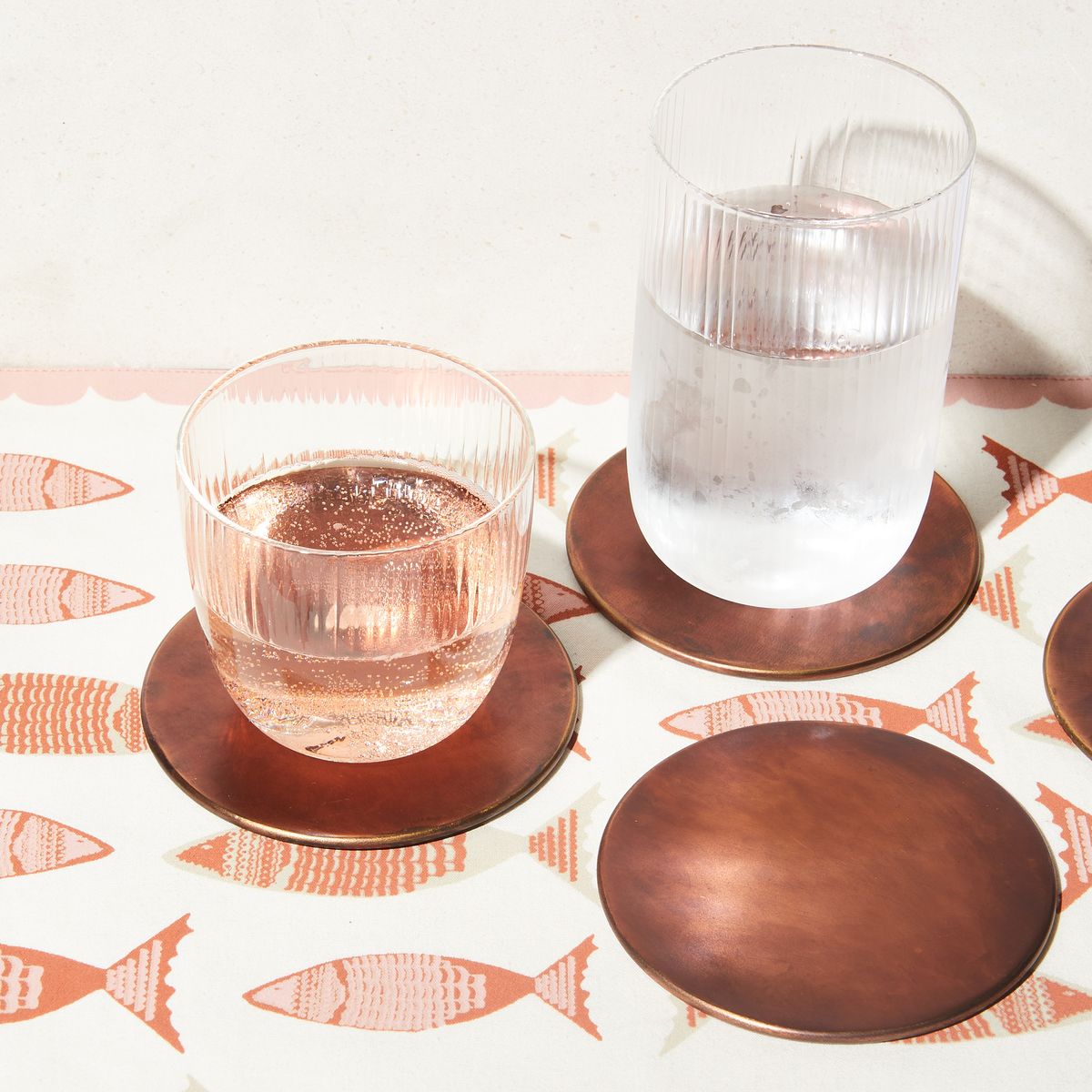 Three copper coaster, one with a short glass, one with a tall glass, on top of a placemat with a fish design