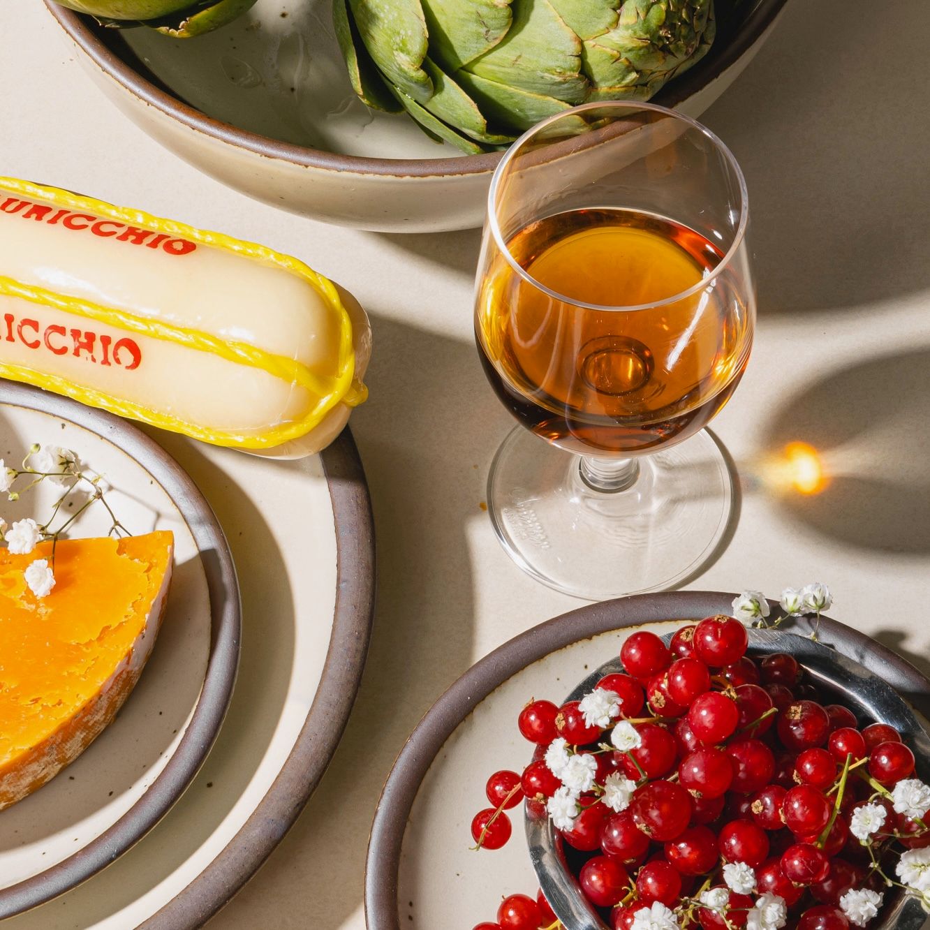 A simple wine glass filled with orange wine sits next to ceramic plates and shallow bowls in a cream color with red berries, artichoke, and a block of cheese.