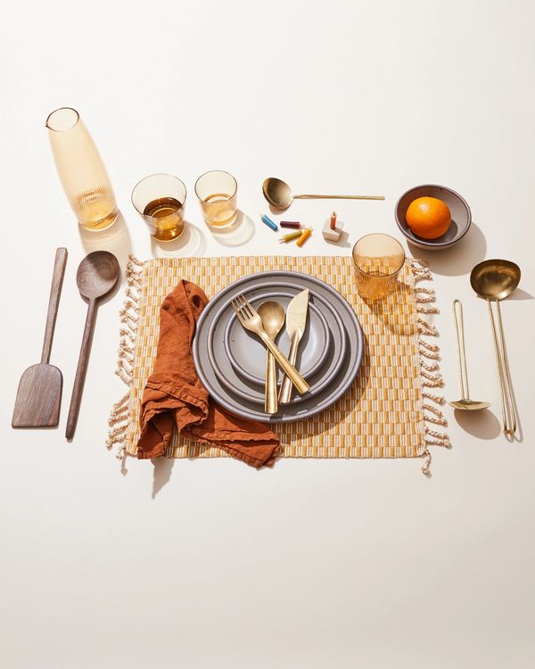 A table setting with a warm placemat, brass flatware, a terracotta napkin, and plates in a warm pale brown, and more.