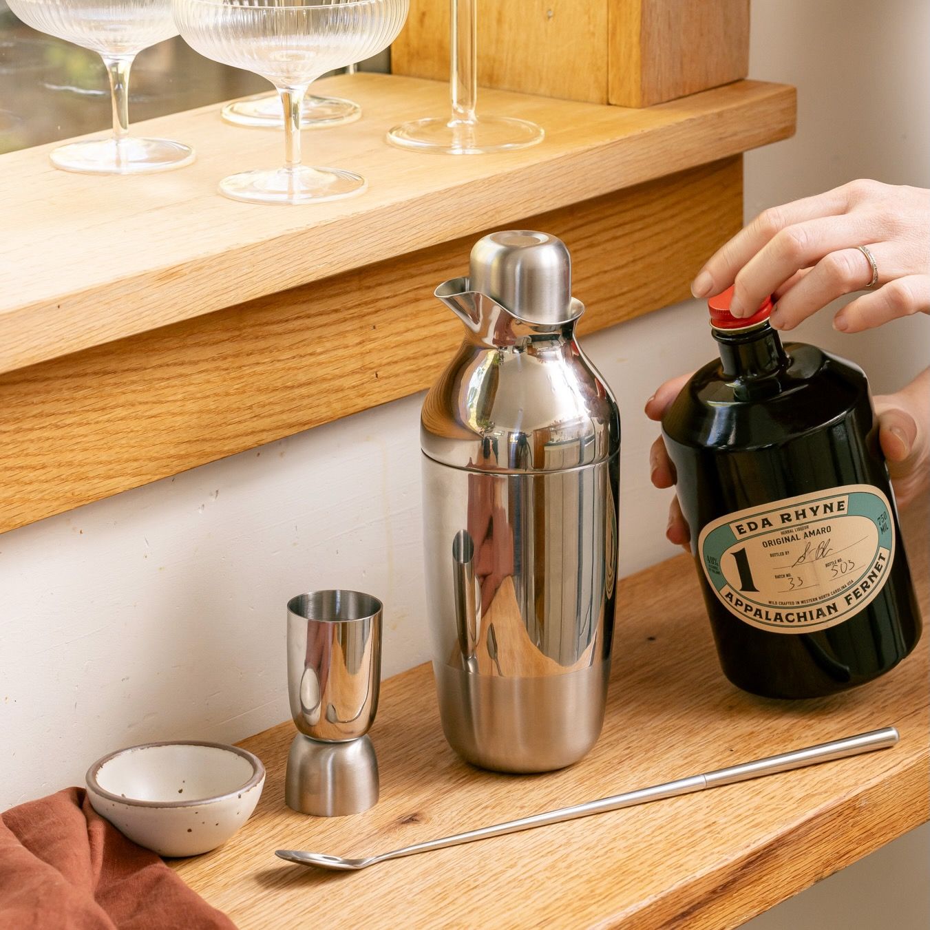 A hand holds a bottle of spirit next to a stainless steel cocktail shaker set on a wooden bar.