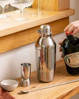 A hand holds a bottle of spirit next to a stainless steel cocktail shaker set on a wooden bar.