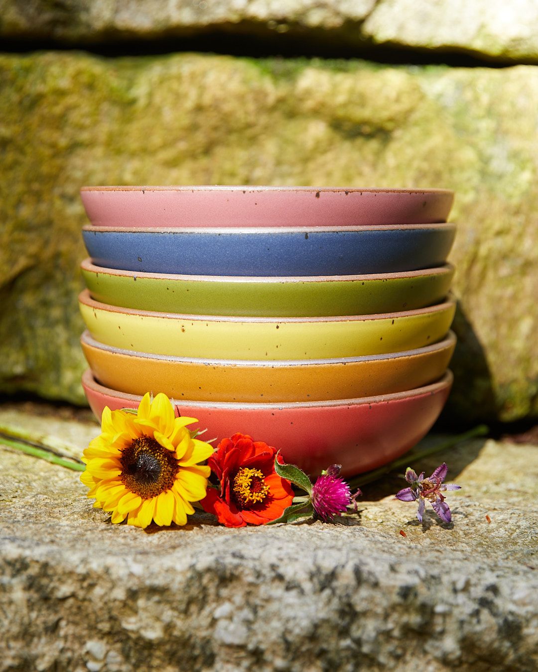 Six bowls in pink, blue, green, yellow, orange, and red sitting on a stone step with flowers in front of them