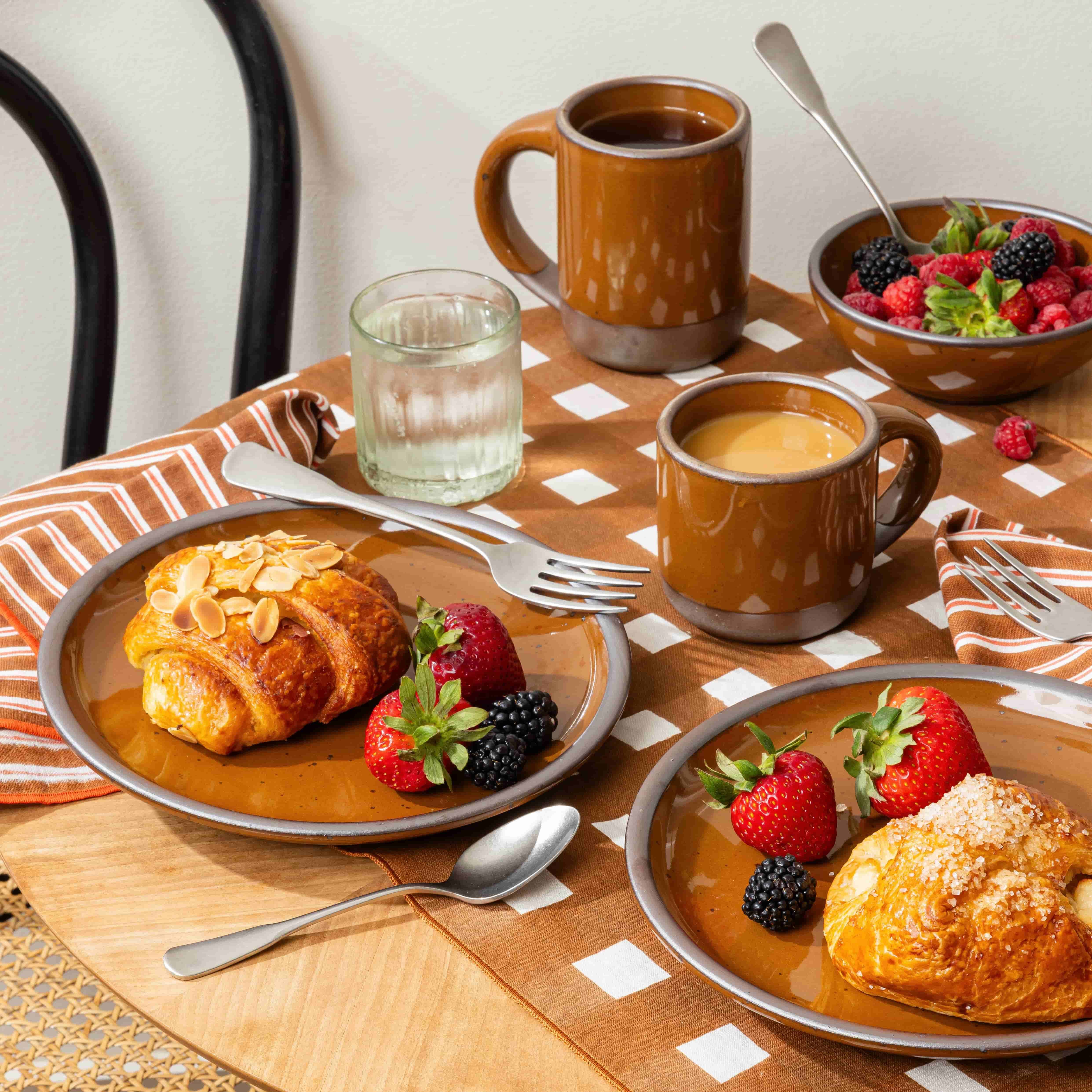 Breakfast table with glossy caramel brown mugs, croissants, strawberries, blackberries, and raspberries on patterned linens.