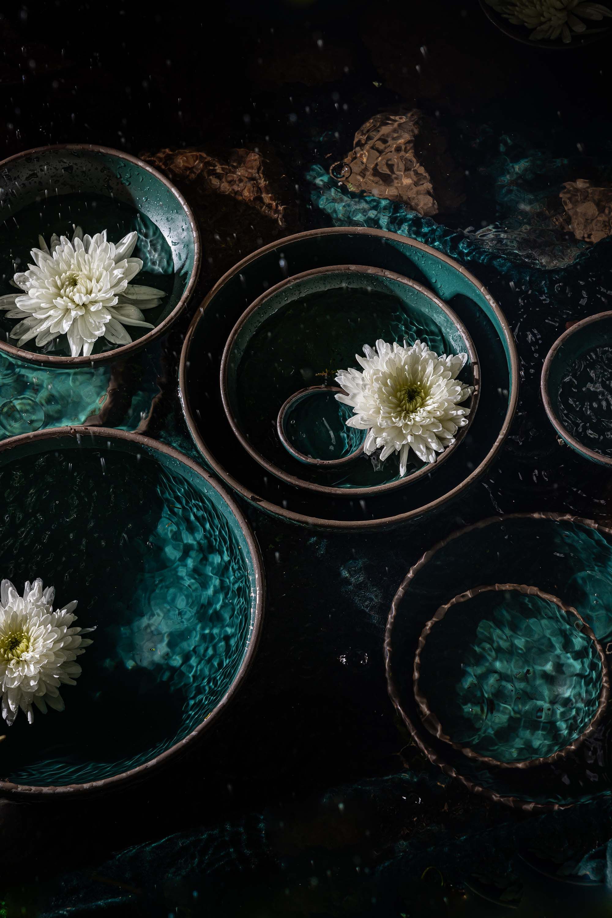 Various plates and bowls in a deep dark teal sitting in dark clear water with white flowers