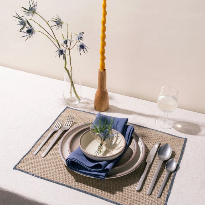 On a table is a place setting featuring a natural woven placemat, ceramic cream plates, steel flatware, a blue linen napkin, and a wine glass, tapered candle, and flowers.