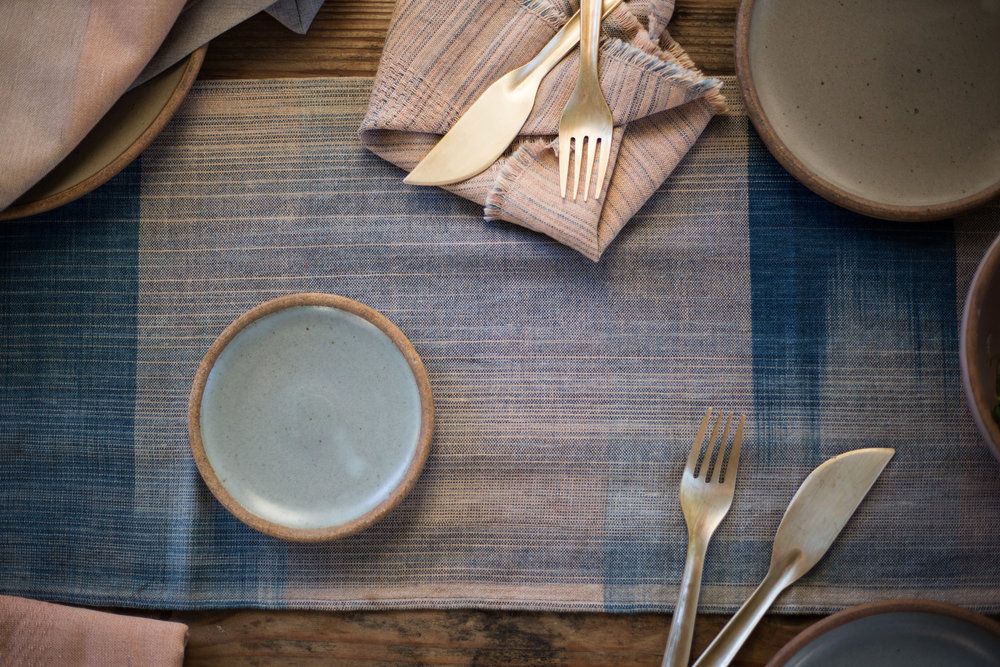 A white and blue woven runner sits on a table with ceramic plates and shiny forks and knives.