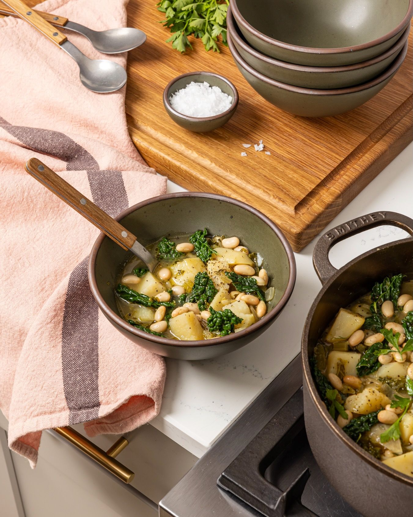 A forest green ceramic bowl filled with a bean soup next to a stove with a petal kitchen towel.