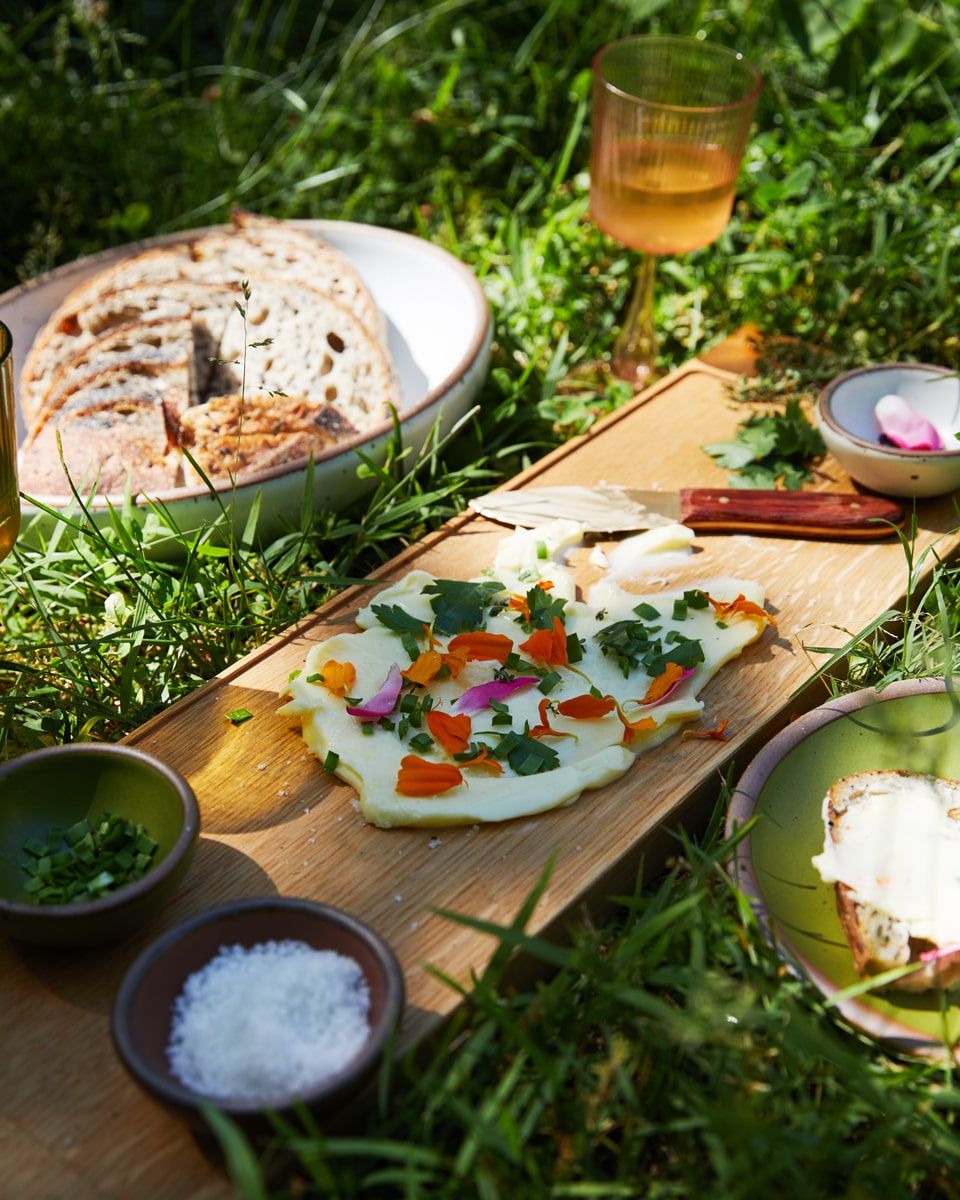 An outside arrangement of a charcuterie board, bowl of fresh sliced bread, butter with beautiful herbs and edible flowers, and glass of wine.