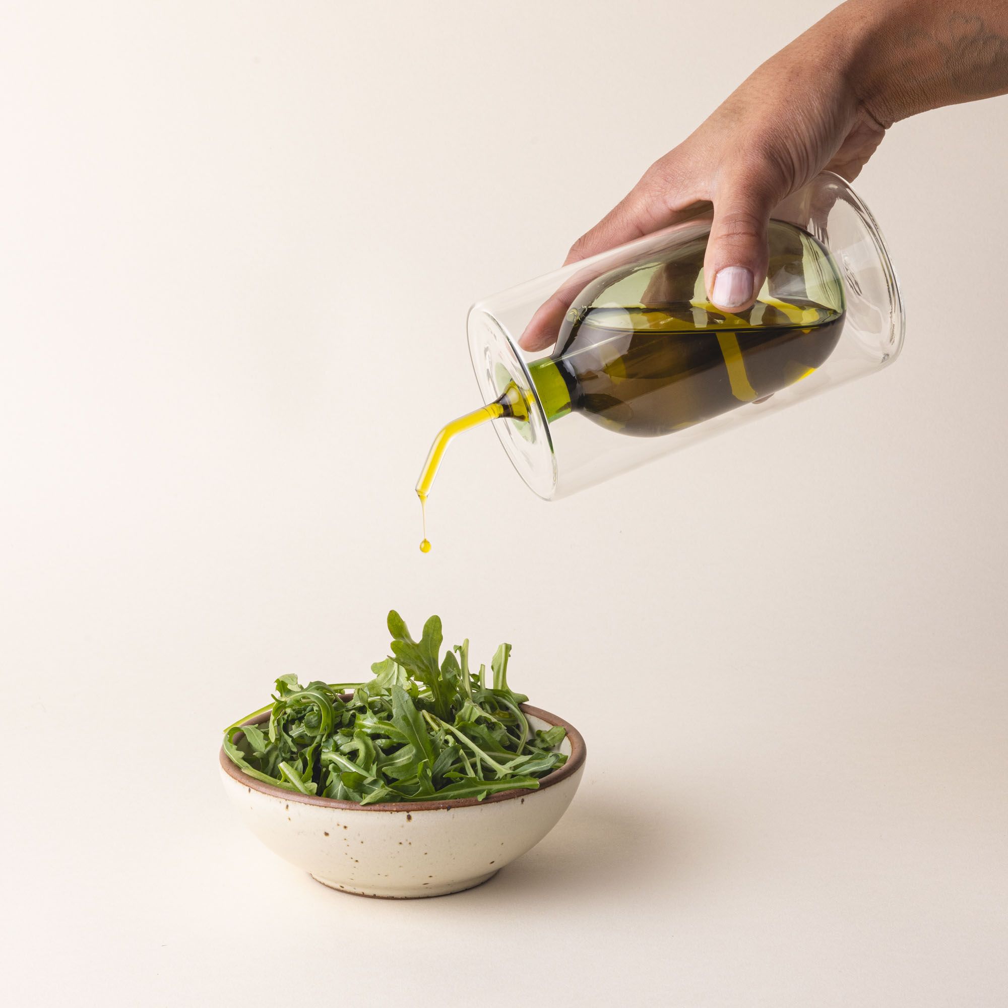 A hand holds an artful glass cruet in a green color and is pouring it over a small side salad. There is a rounded bulb that acts as a container for the cruet, surrounded by a doubled glass wall. The cruet is filled with oil.