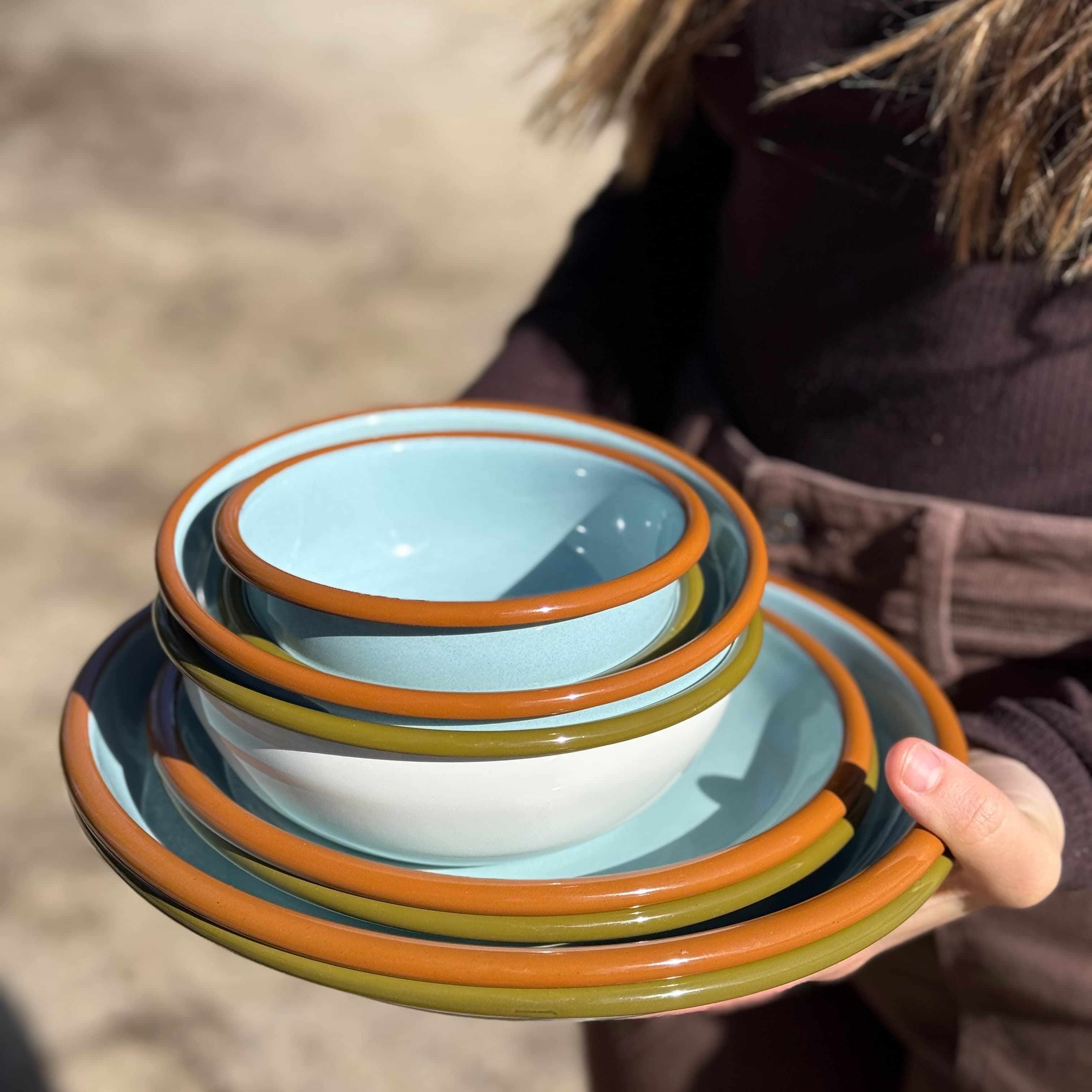 A woman holding enamel plates and bowls stacked together in robin blue and white colors with olive green and warm brown rims.