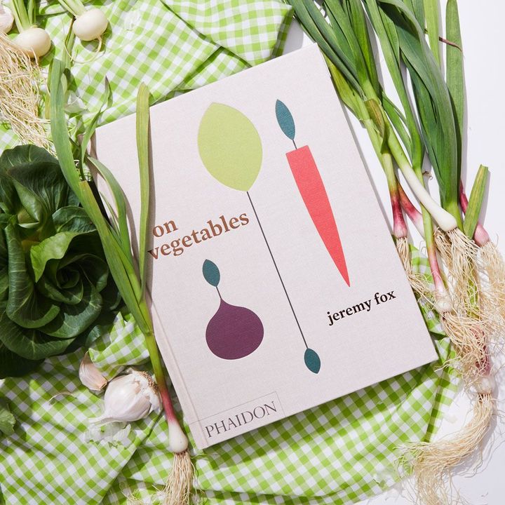 White cookbook with simple graphics of vegetables on the cover; on a green gingham picnic blanket with an assortment of green vegetables framing it