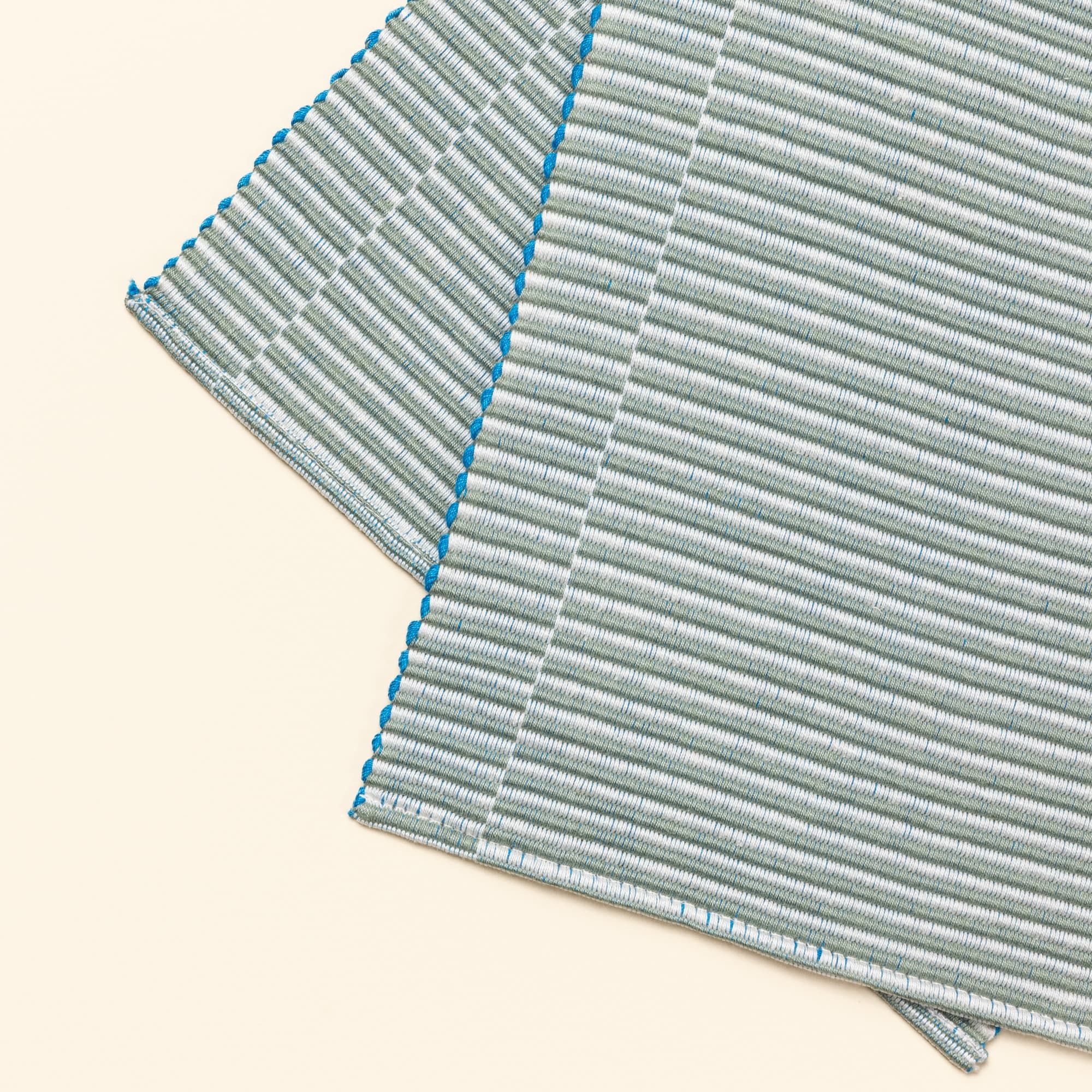 A close up of a corner of a table runner with offset stripe design that is hand woven in sage and blue colors.