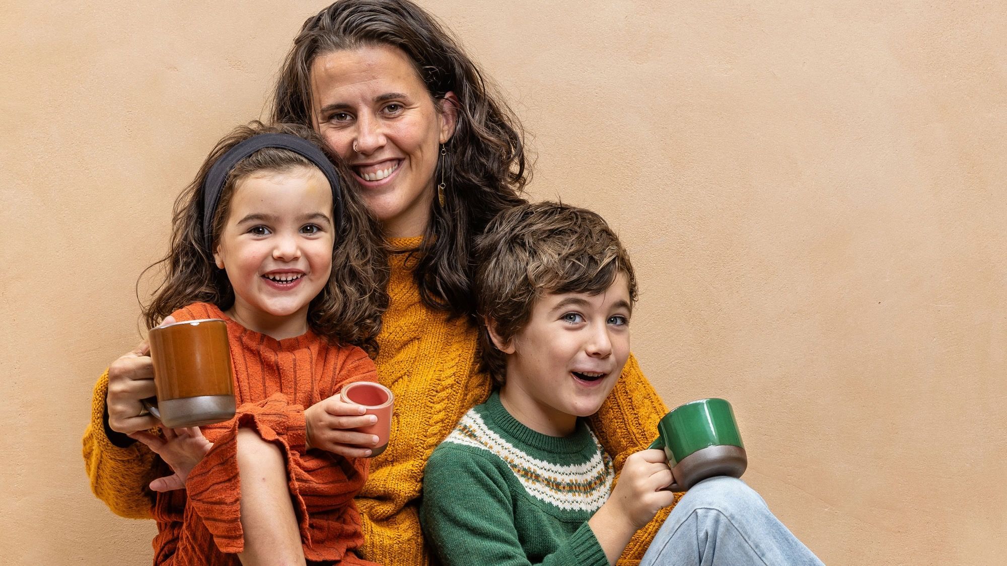 Patty smiling with her two kids, and each of them are holding a ceramic mug or cup in glossy brown, glossy green, or pink colors.