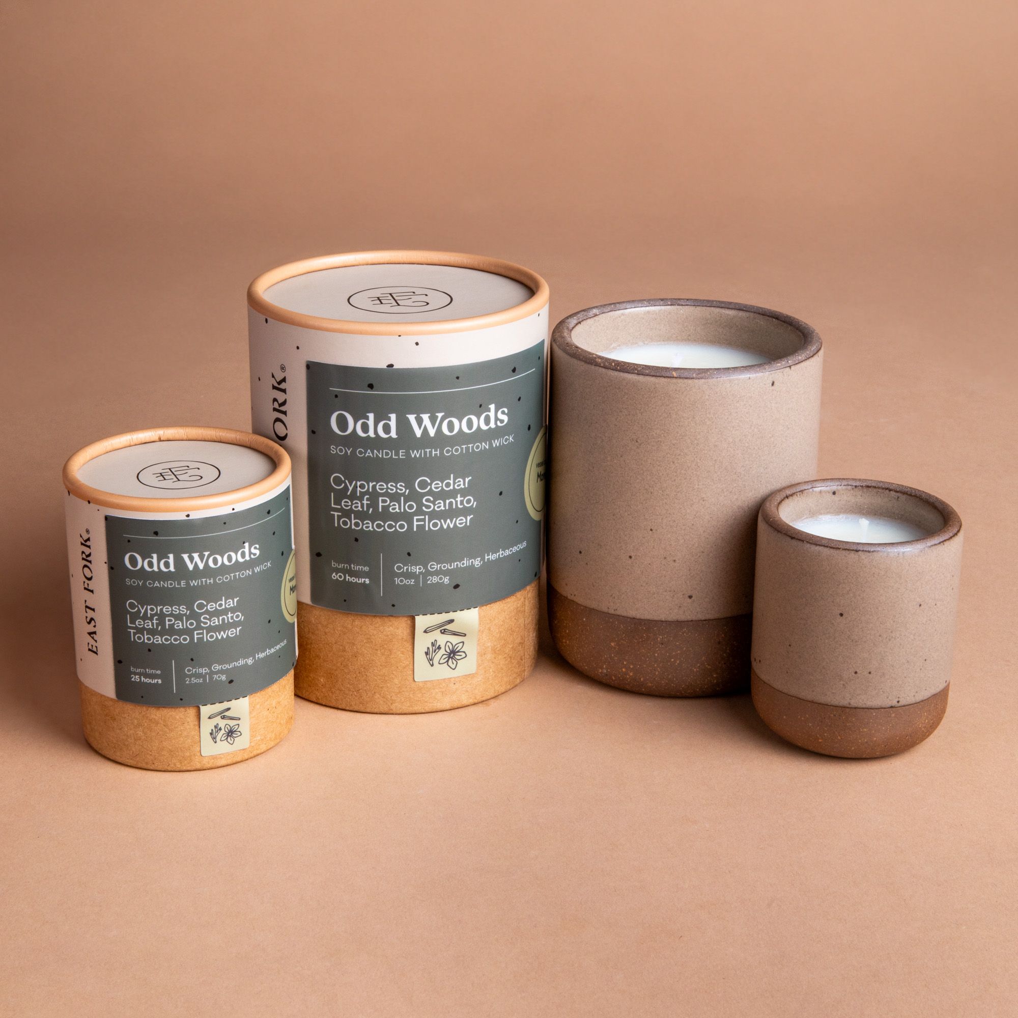 Small and large ceramic vessel next to each other in a warm pale brown color with candles inside each. Cardboard tube packaging is on the left with branding stickers that say "Odd Woods".