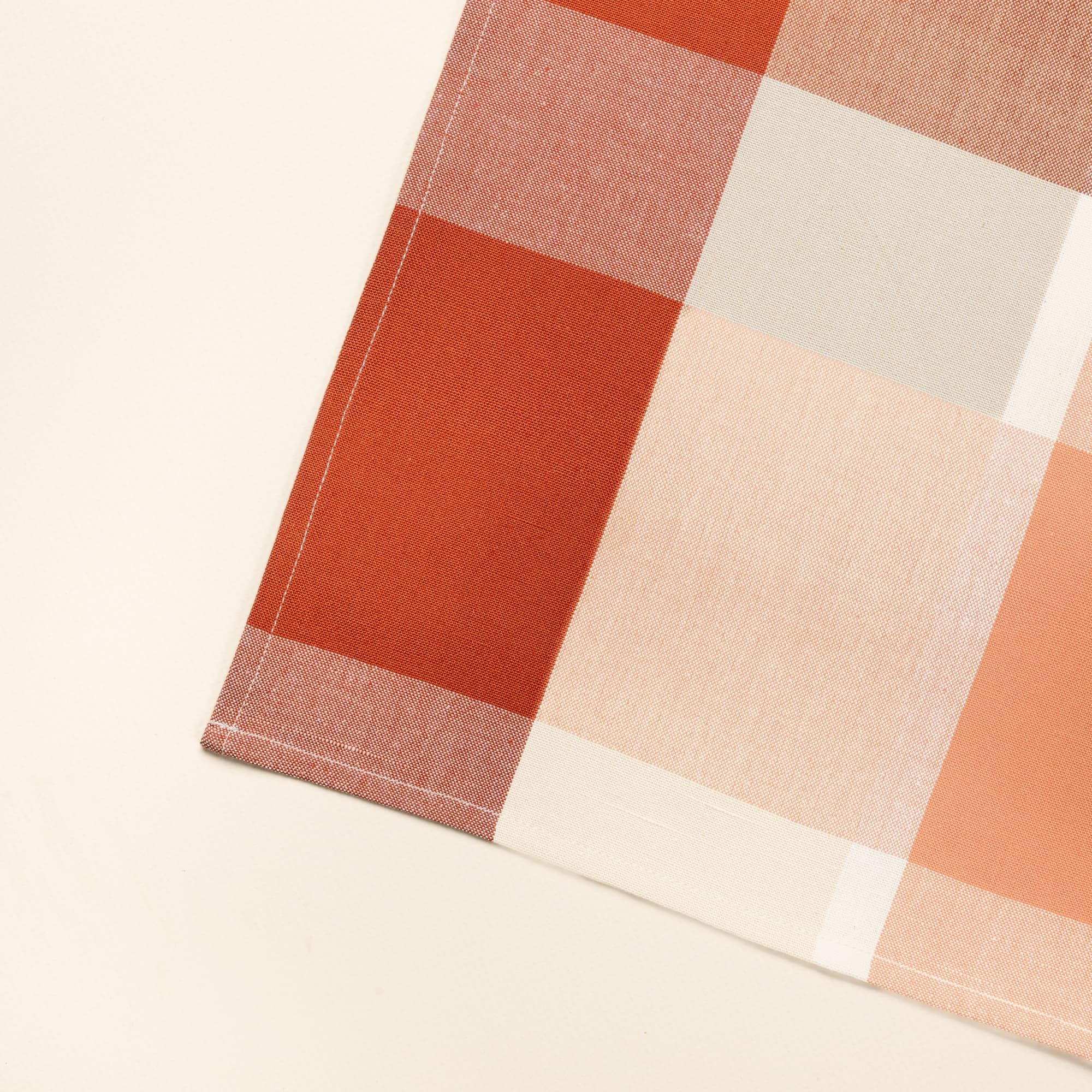 A close up of a corner of a flat square dinner napkin in a rust and cream gingham pattern.