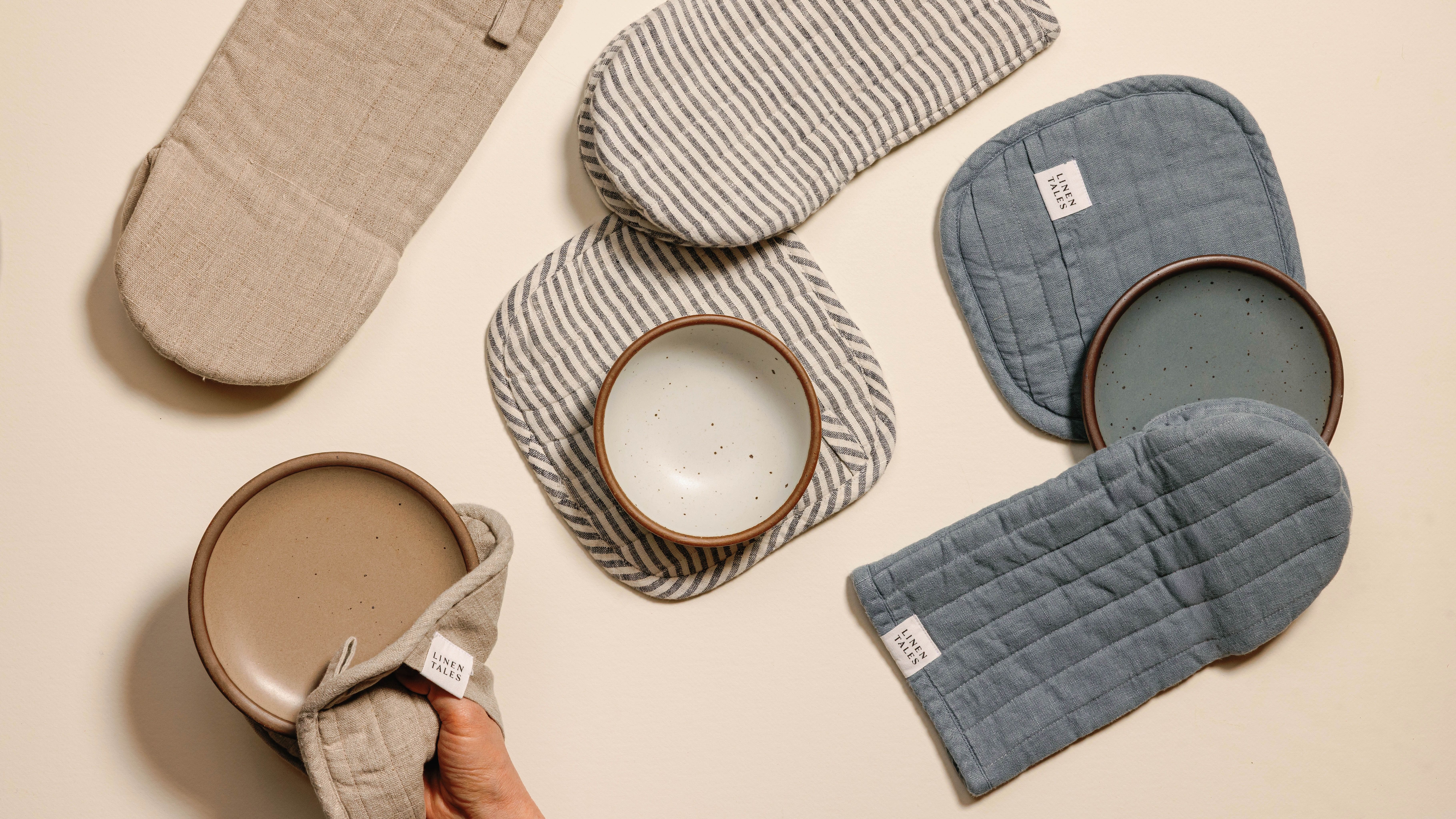 Linen oven mitts and pot holders in natural, striped, and blue-grey colors with small ceramic plates in the same colors.