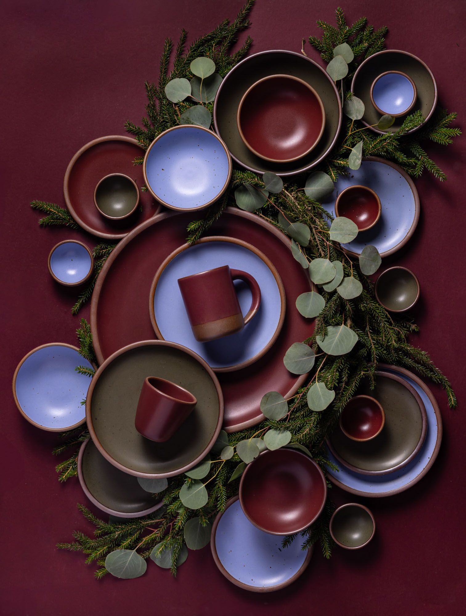 Ceramic plates, bowls, and mugs in a forest green, periwinkle, and deep plum red artfully laid with festive greenery.