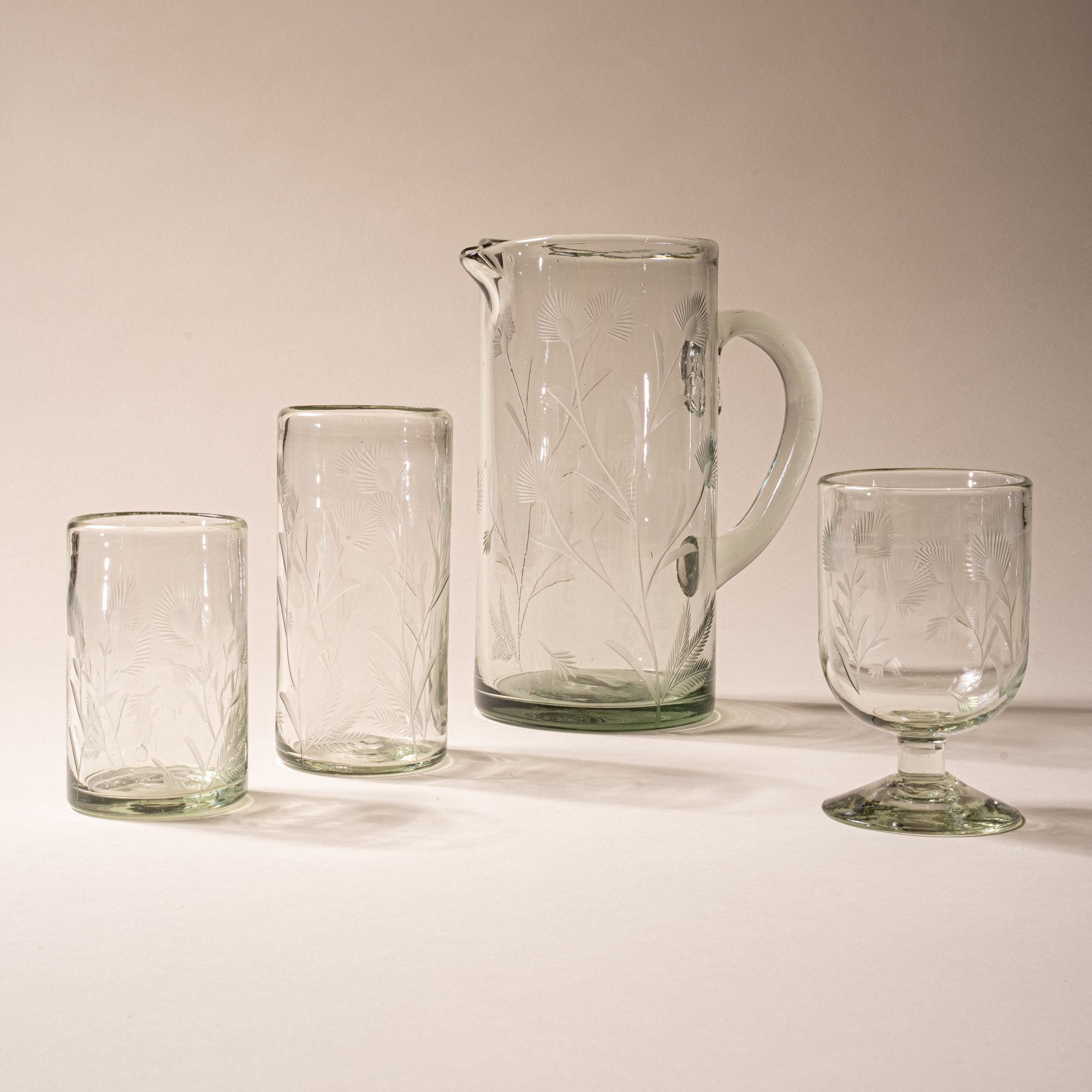 Set of clear etched glassware with floral design, including pitcher, wine glass, highball glass, and short tumbler on neutral background.
