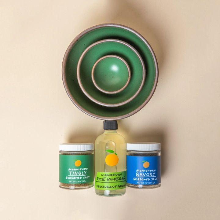 3 Ceramic bowls of varying sizes nested together in a cool green color with 2 jars of Momofuku seasoned salt and a bottle of rice vinegar below.