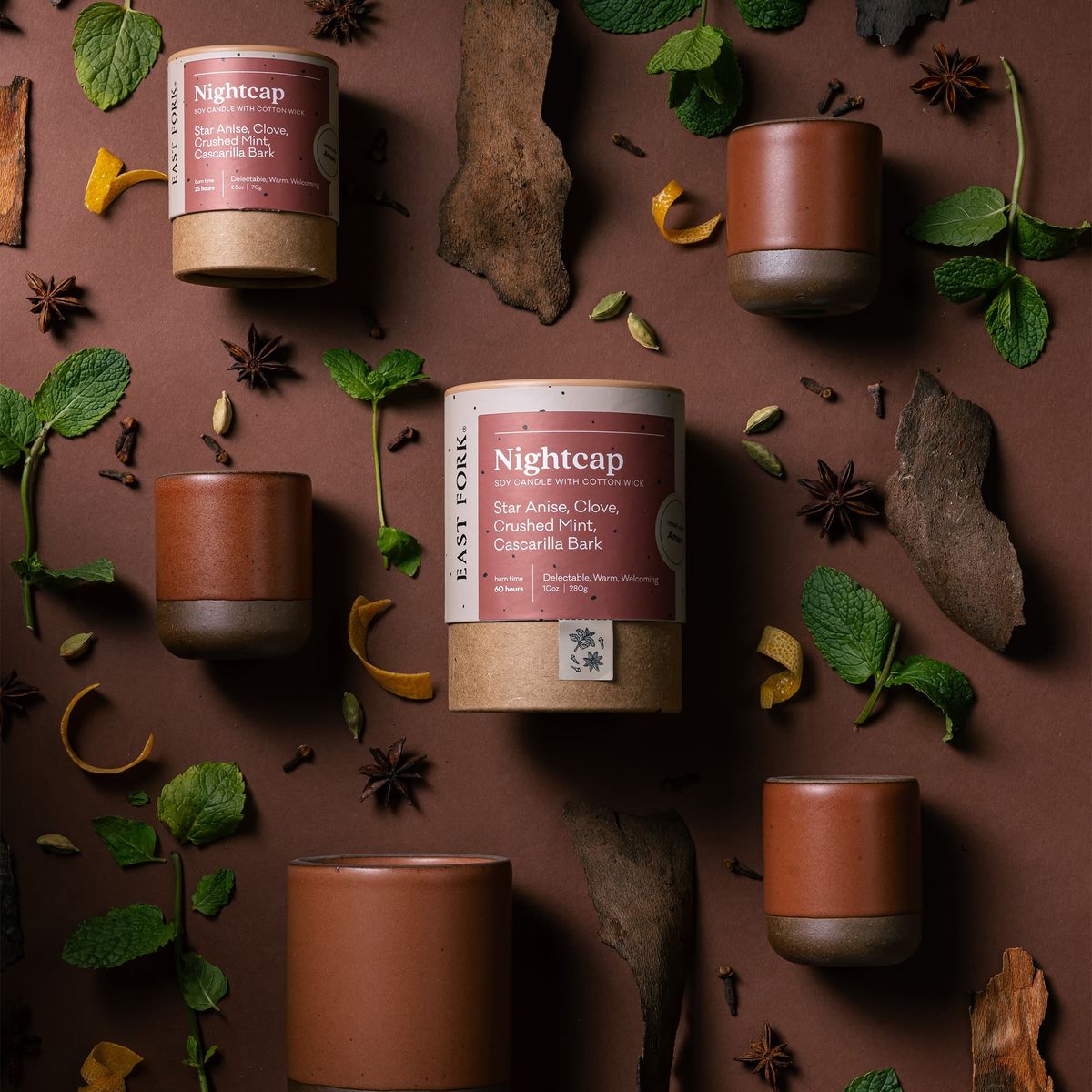 An artful layout of The Candle in Nightcap - centered is a cardboard packaging tube with the candle's name, surrounded by small and large ceramic candles in a cylindrical vessel in a cool terracotta color, surrounded by anise, mint, clove, and bark.