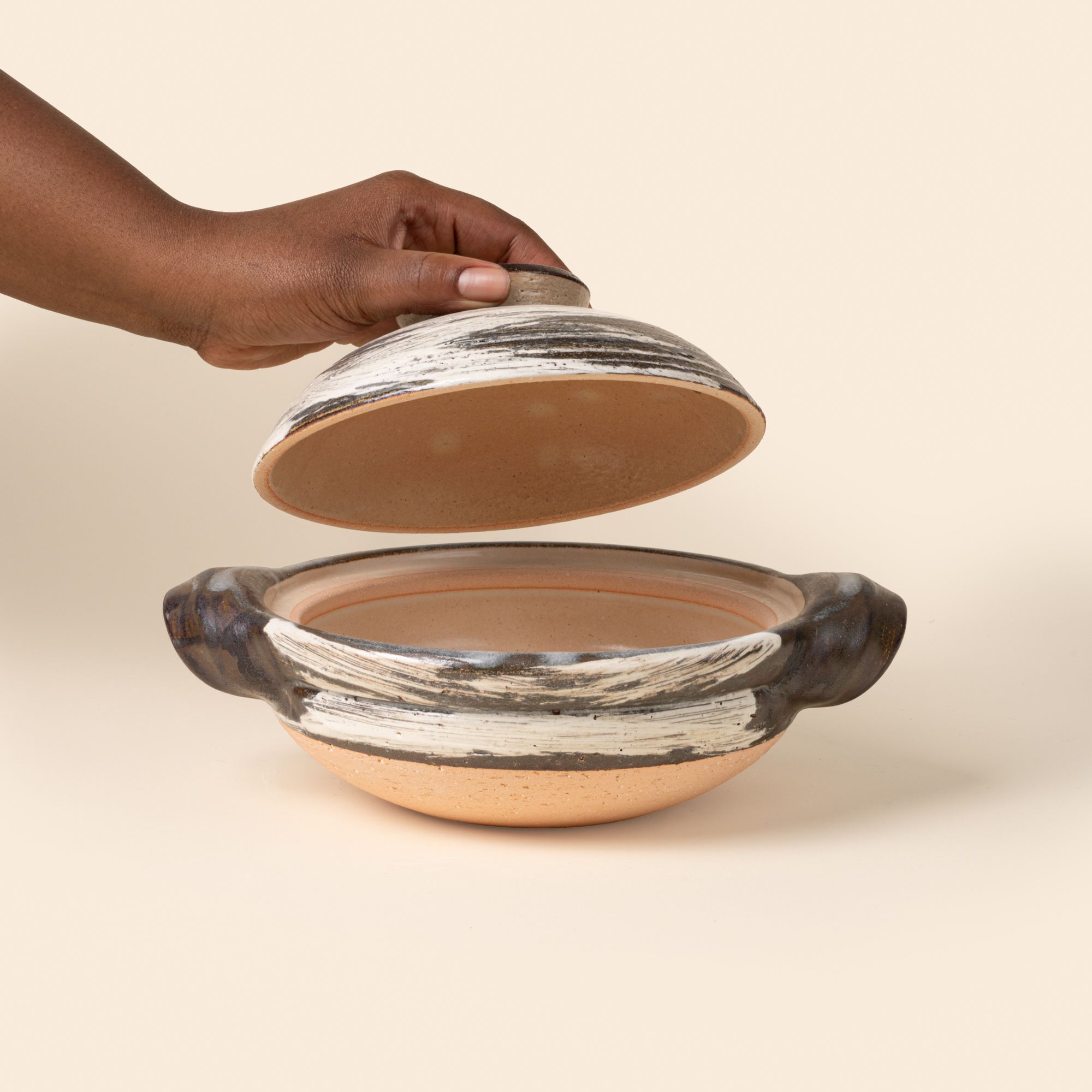 A hand holds the lid over a ceramic donabe with side handles, finished in a black-and-white brushed glaze over natural clay.