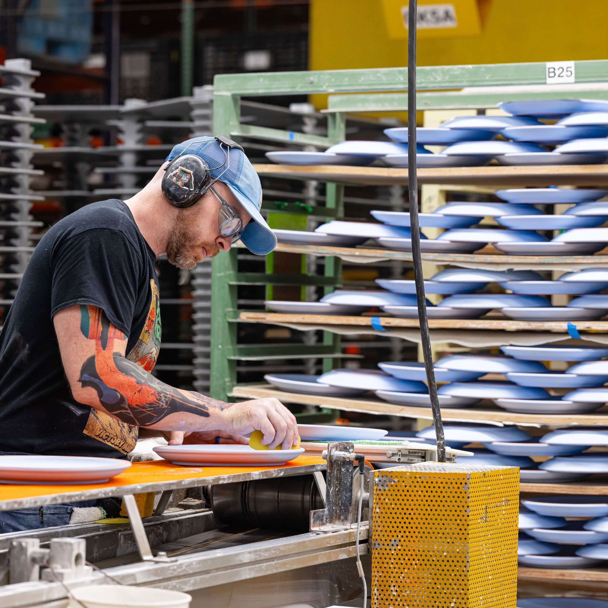 In a factory setting, a person is working on ceramic plates and there are large shelves holding periwinkle ceramic plates