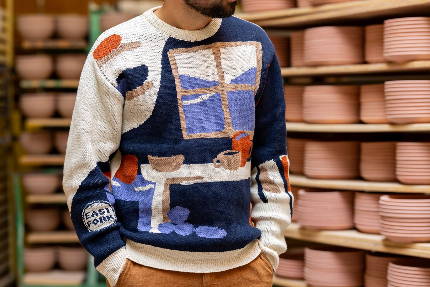 Alex wears a sweater with an illustration of a pottery workshop with a large window, shelves, and table with mugs and bowls. Alex stands in front of a rack of ceramic bowls in progress.